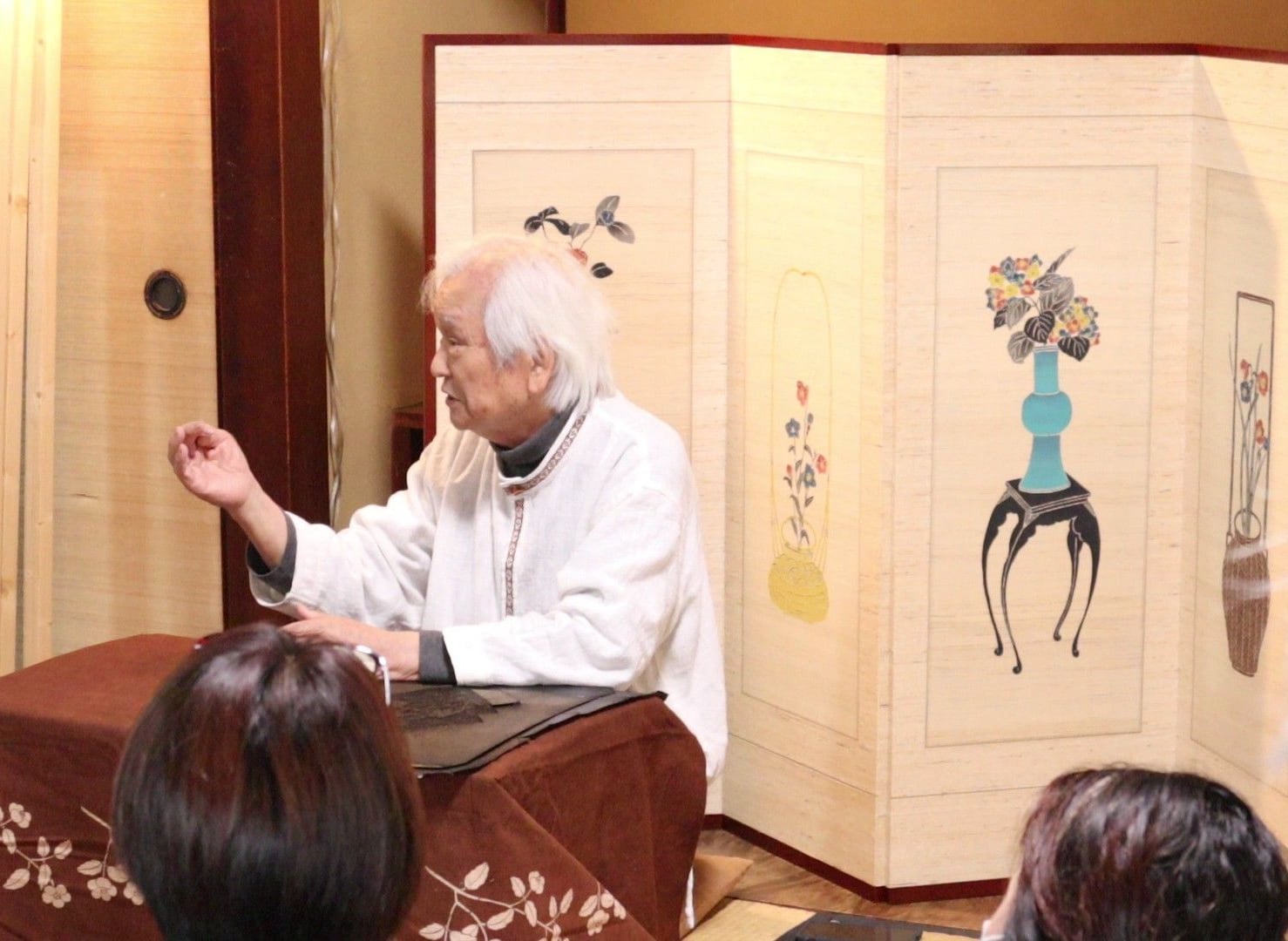 和室で、花器を描いた屏風を背に話す下平先生。手前に参加者の後ろ姿が見える
