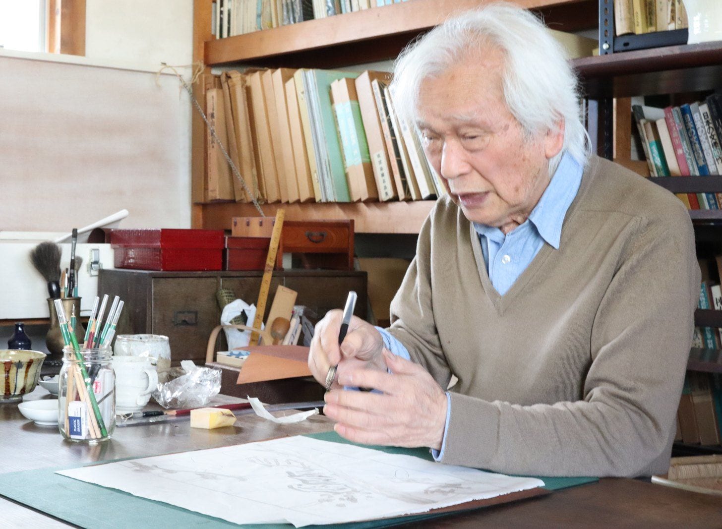書棚の前の机に向かい、図案に手を入れる下平清人先生