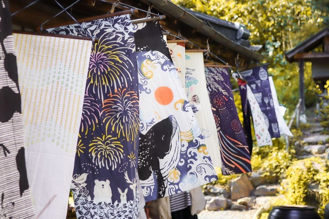 Patterned tenugui hand towels hung in a row under the eaves of a wooden house, with greenery behind