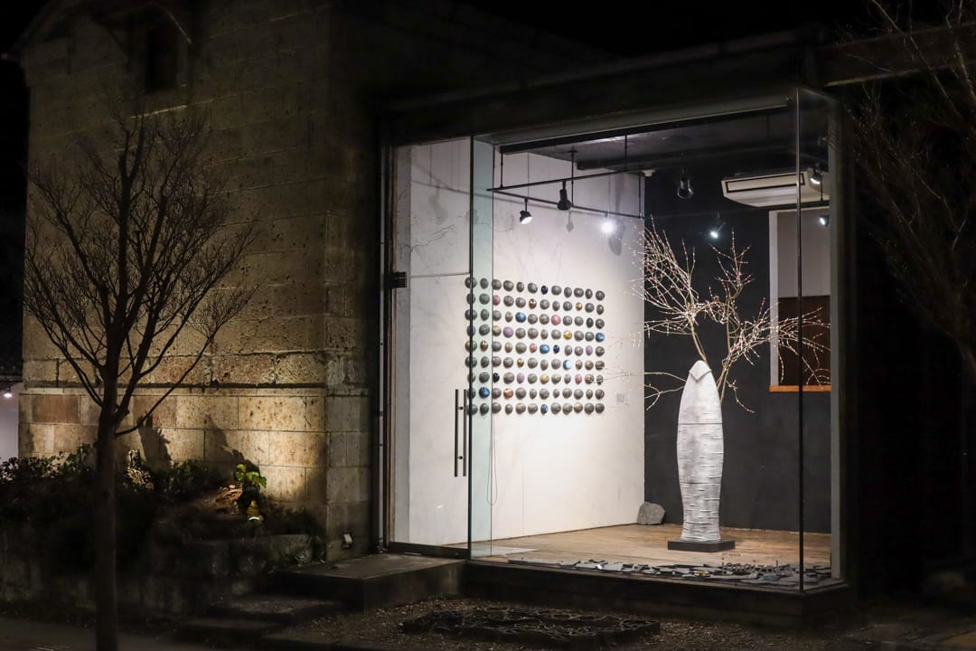 Art Space Jonaizaka at night: a glass-fronted white cube beside the stone wall, lit from inside with an installation of a white vessel and branches