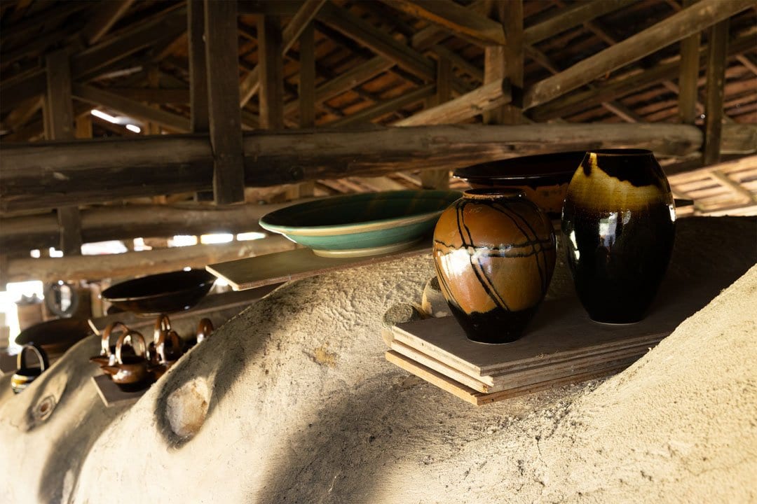 Glazed jars and a green dish resting on the clay ledge of a kiln beneath a timber roof