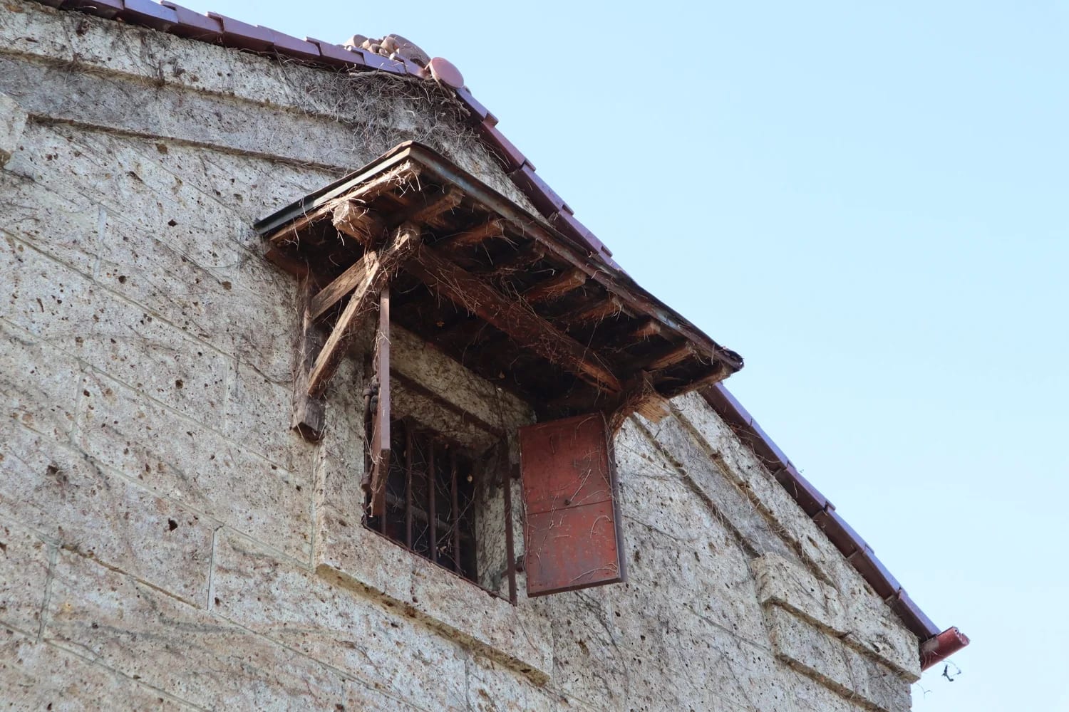 Close-up of the rough Oya stone wall of Toko’s warehouse, with a small wooden window under the tiled eave against a pale sky