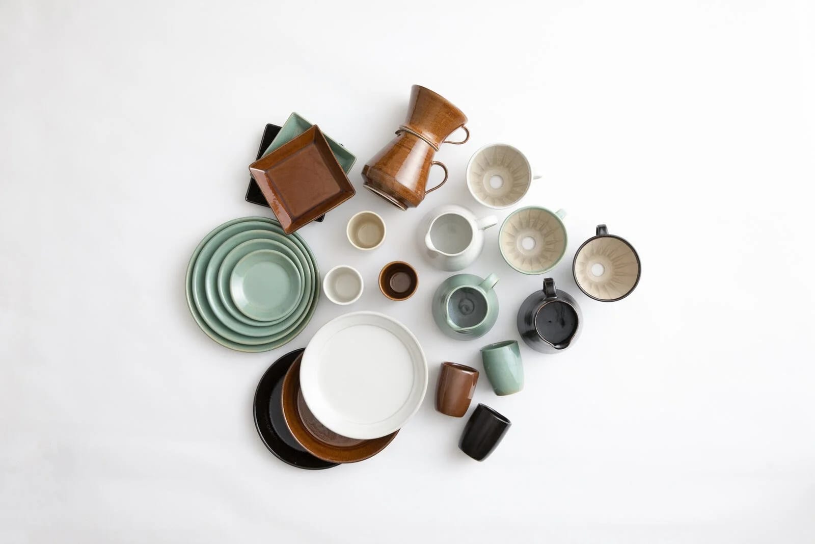 Plates, bowls, cups and a small pitcher by Sayado Wadagama in muted green, white, brown and black glazes, laid out on a white surface
